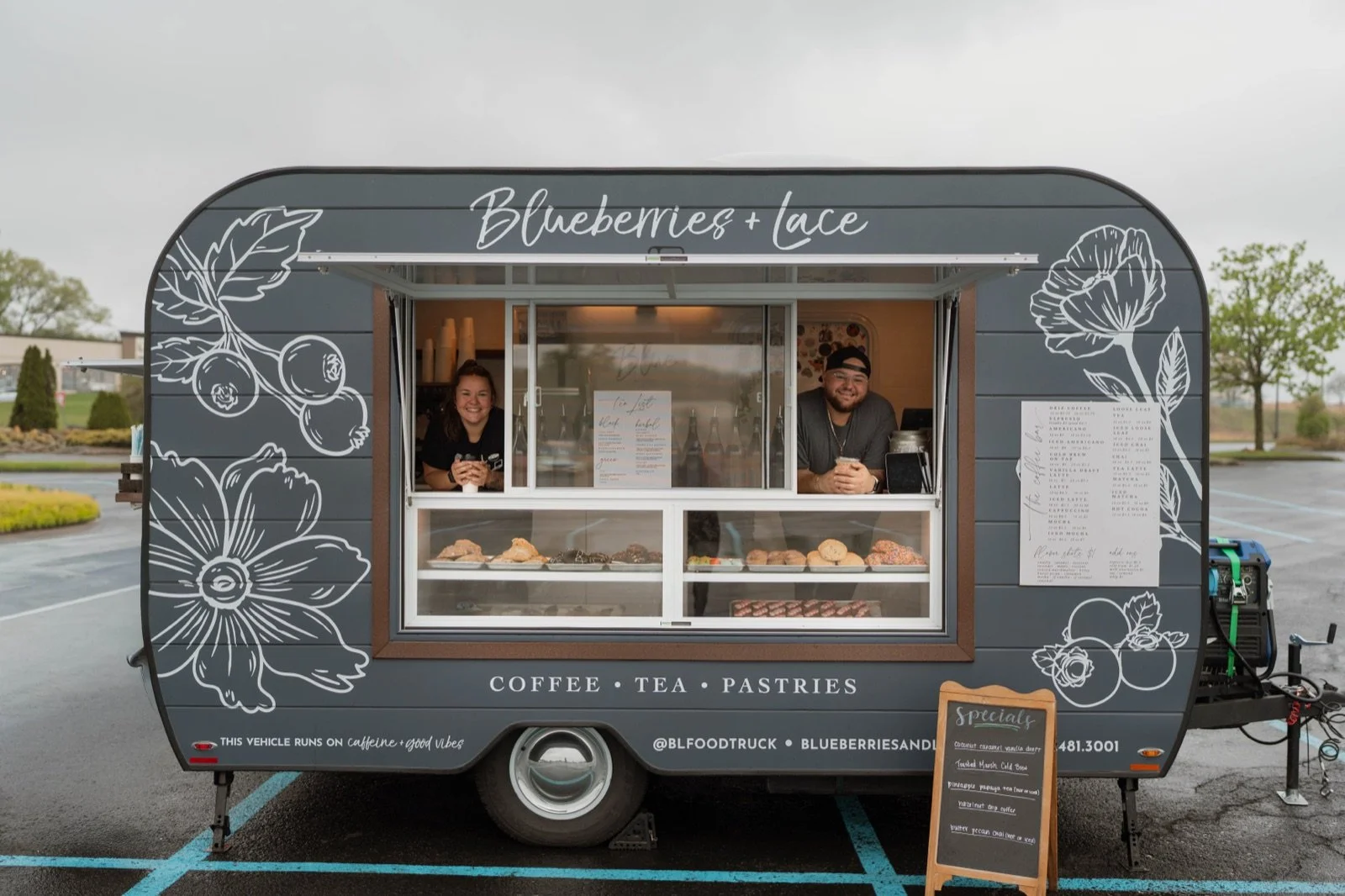 Blueberries & Lace’s gray Aero Build coffee trailer with floral branding, an open service window, and two people serving drinks.