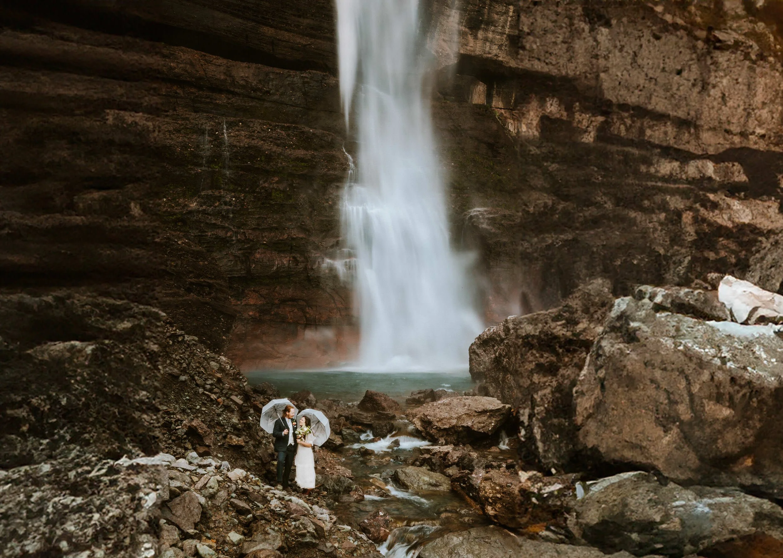 A couple in wedding clothes stands under bridal veil falls in Telluride, Colorado while carrying flowers and clear umbrellas