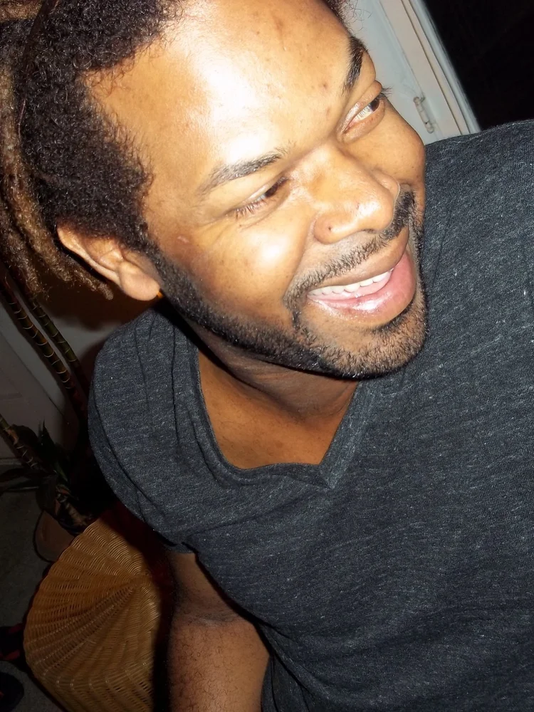 Shah Smoak is a Black person with black hair, beard, and goatee wearing a black t-shirt, smiling and looking to the side. 