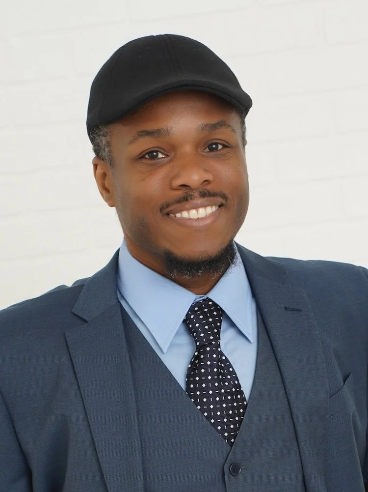 Skylar Richardson is a Black person with a black mustache and goatee wearing a black hat, blue suit jack jacket and vest, a blue shirt and black polkadot tie. 