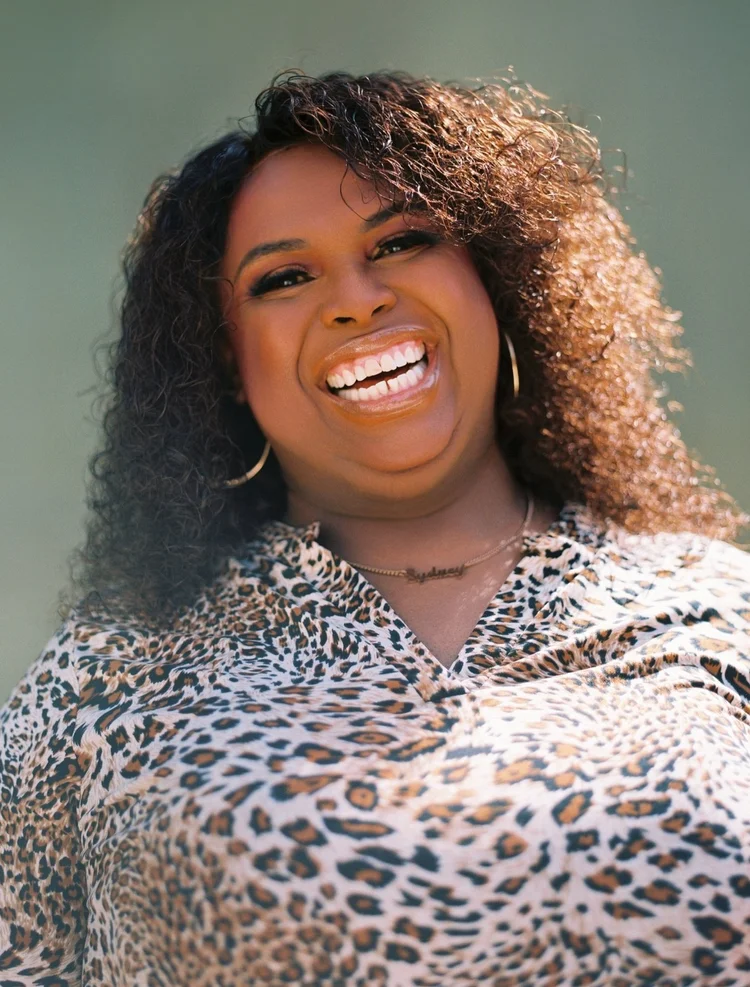 Sydney Rogers is a Black person with long curly brown hair smiling, wearing a leopard print blouse, a necklace, and gold earrings. 