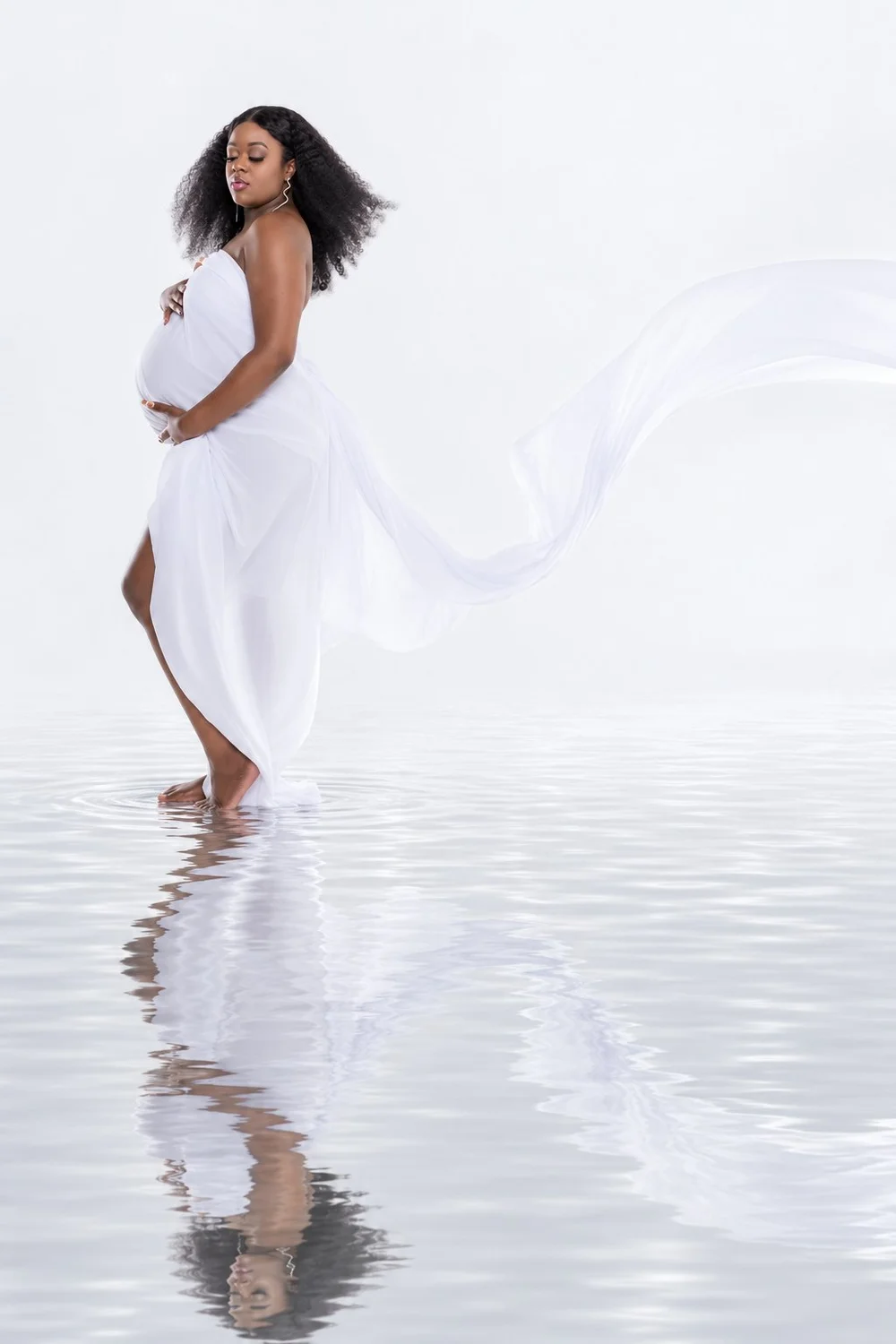 Maternity portrait of Capria cradling her belly in white fabric with a water reflection