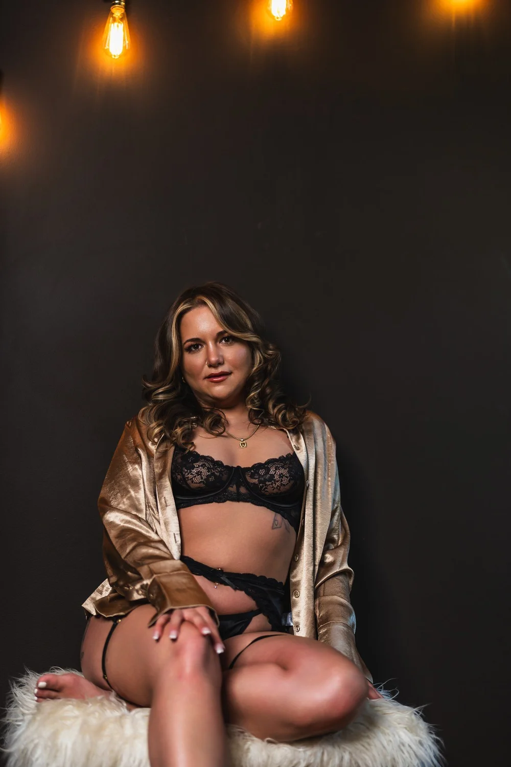 Alicia seated in a satin robe and black lace beneath warm pendant lights