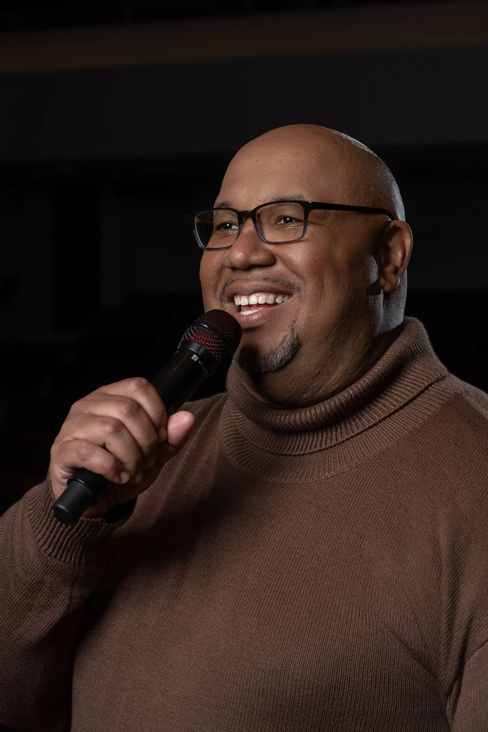 Smiling close-up of Dr. Michael Dorsey in a brown turtleneck holding a microphone