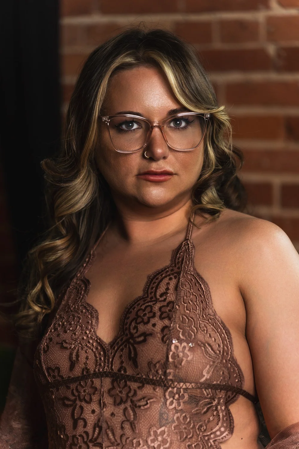 Close portrait of Alicia wearing glasses and brown lace against exposed brick