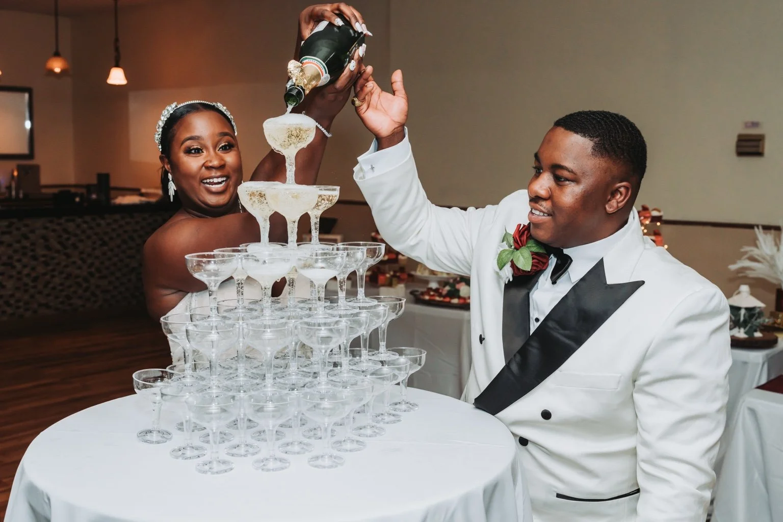 Bride and groom pouring champagne into a tower of coupe glasses at their wedding reception.