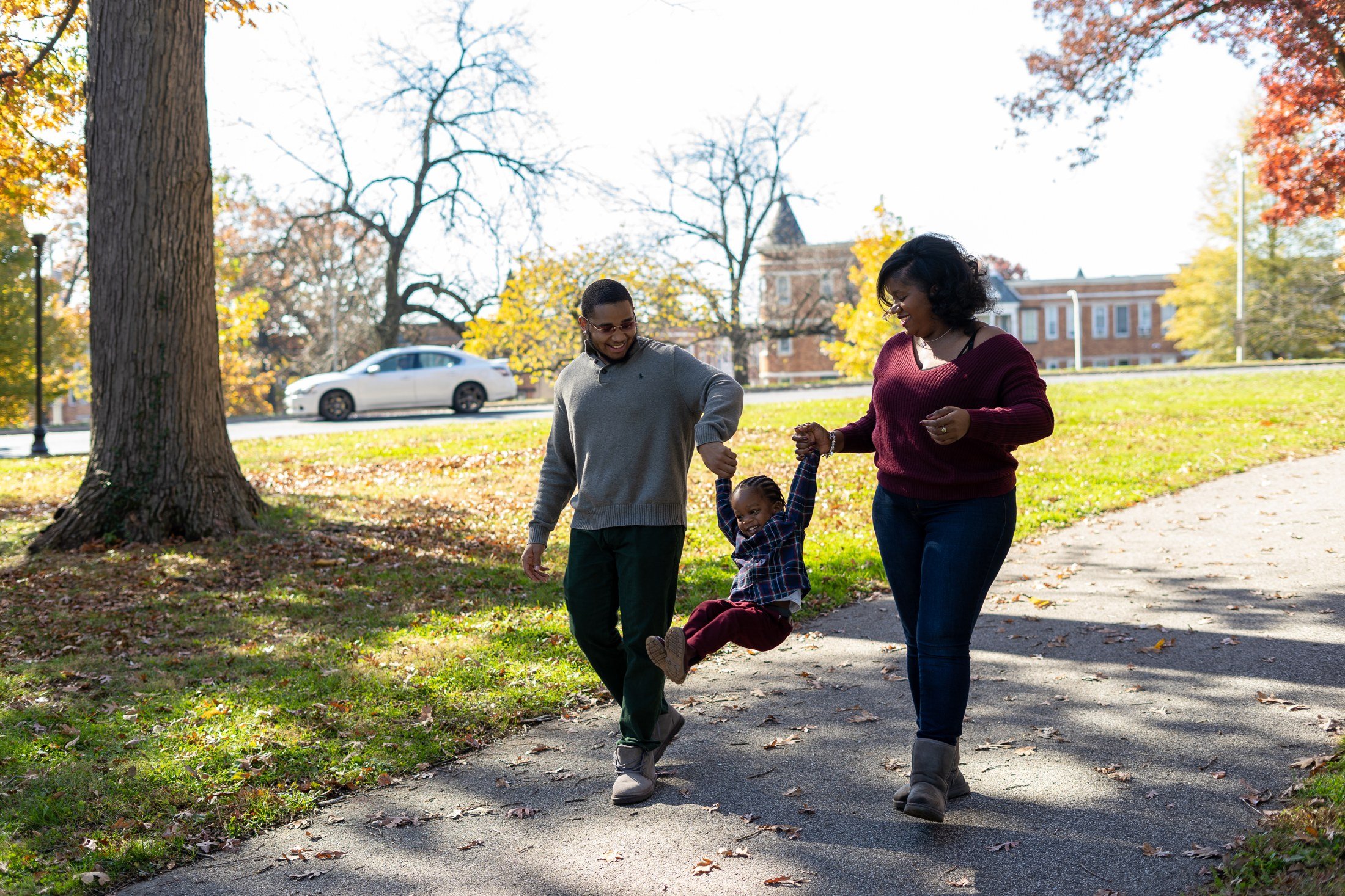 Two adults laughing as they lift a child between them on an autumn park path