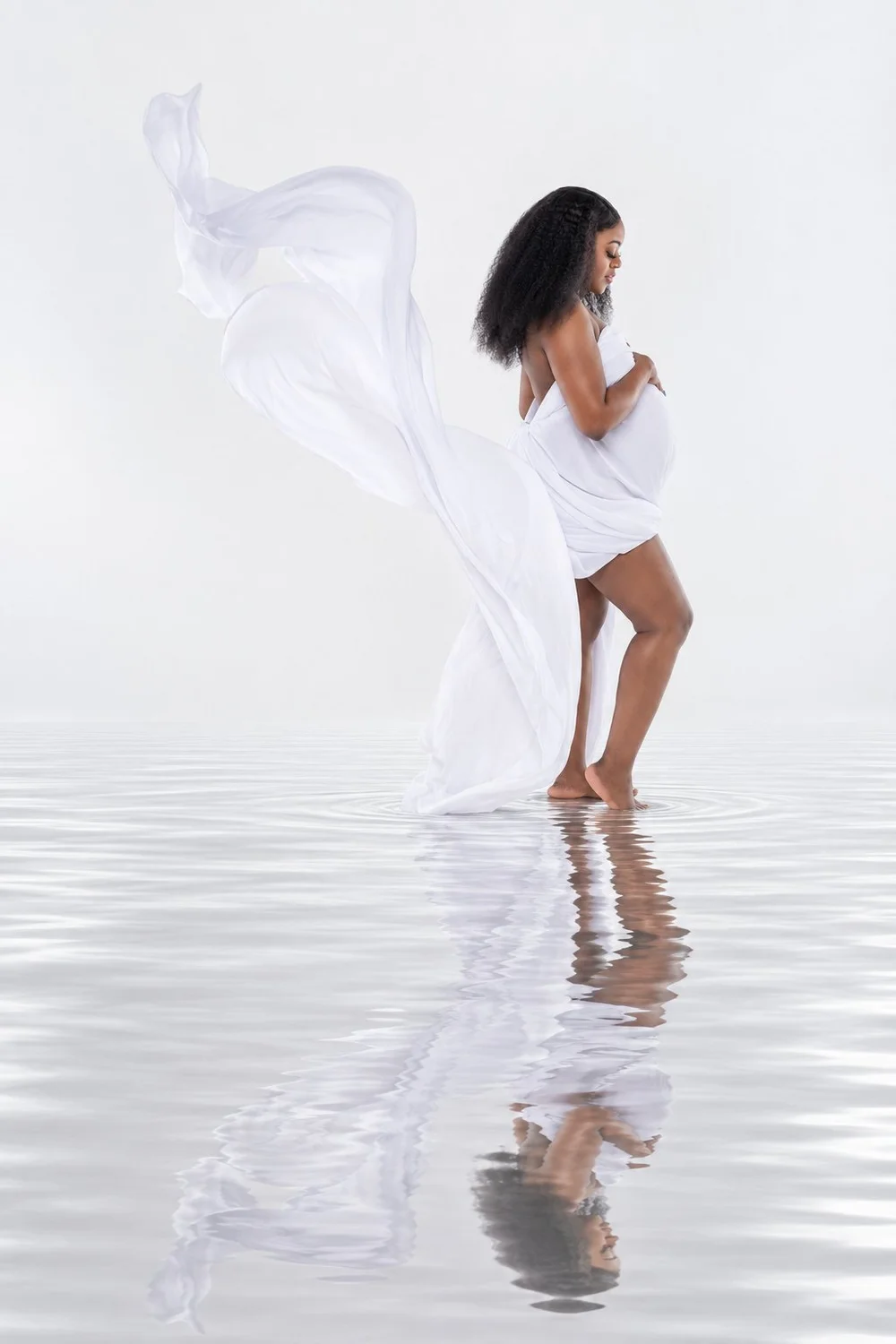 Side-profile maternity portrait of Capria with billowing white fabric and reflective water