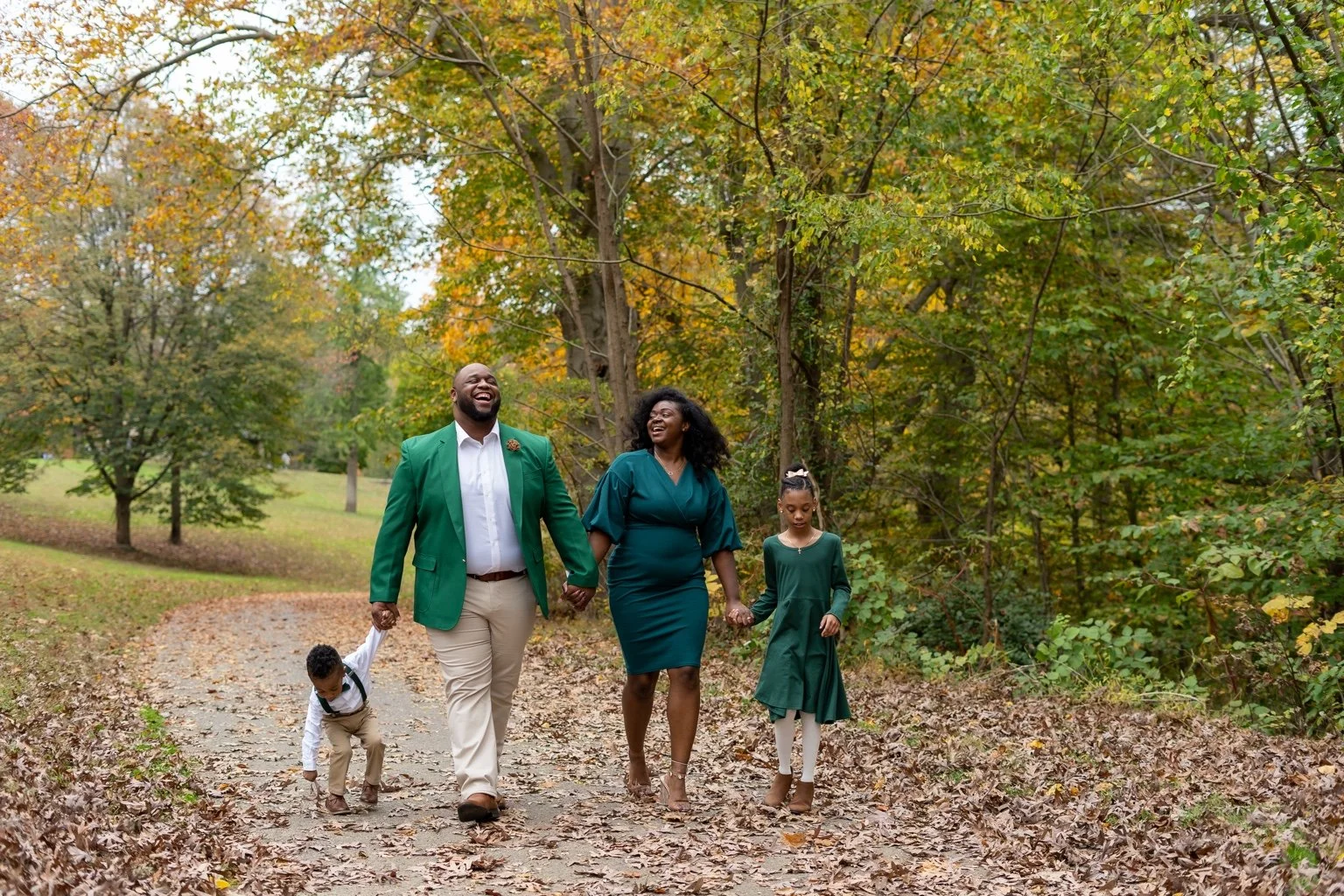 A family in coordinated green outfits laughing and walking together along a leaf-covered path