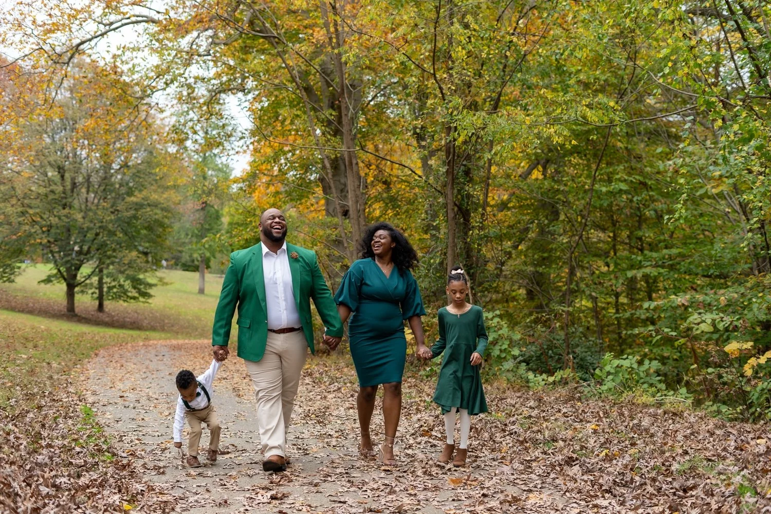 Family of four in coordinated green outfits walking hand in hand along a leaf-covered autumn path