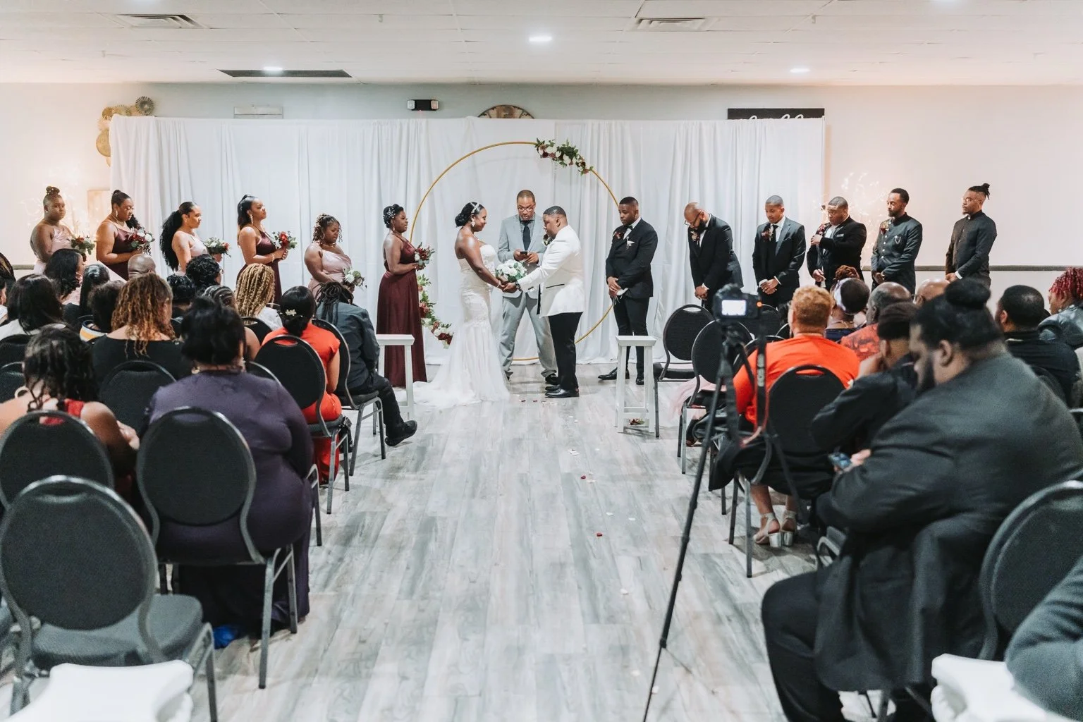 Bride and groom holding hands beneath a circular floral arch, with their wedding party and seated guests surrounding them.