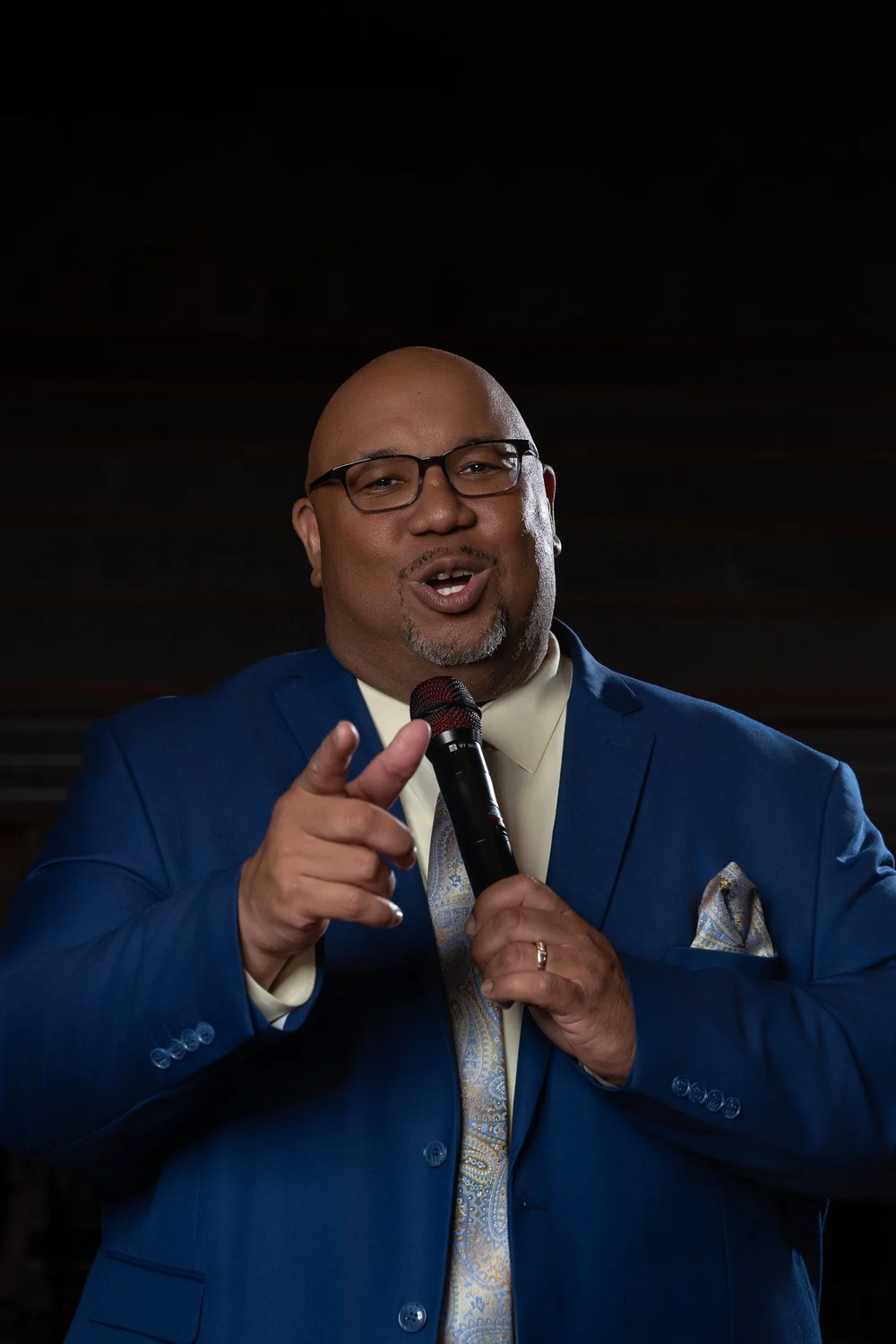 Dr. Michael Dorsey pointing toward the camera while holding a microphone in a blue suit