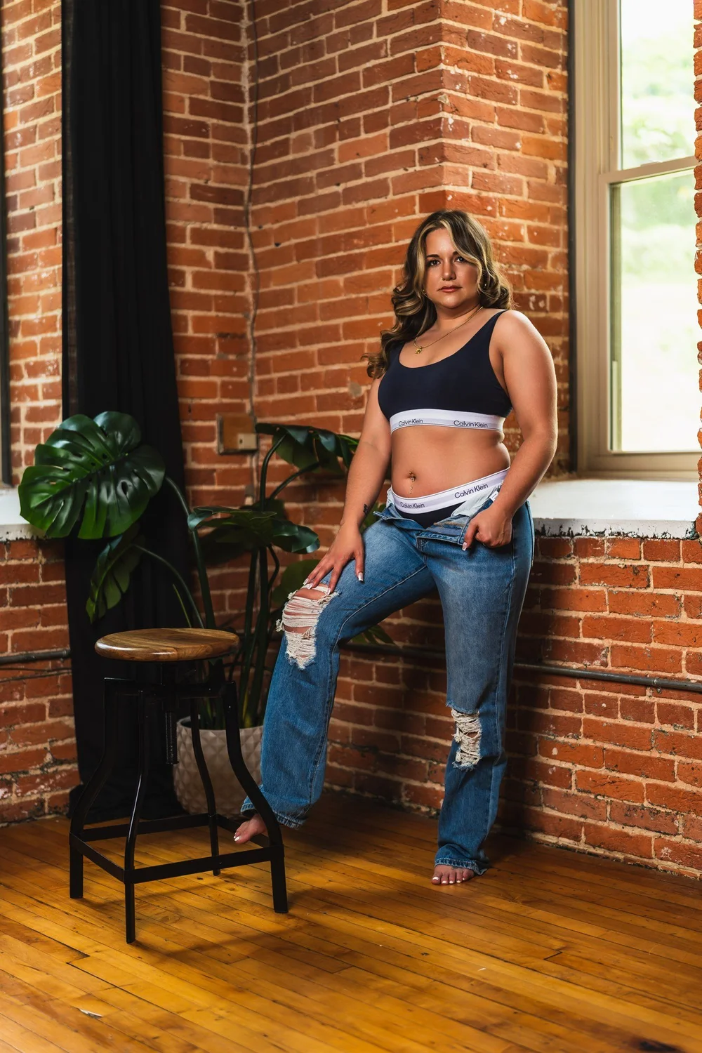 Alicia in a black crop top and denim beside a sunlit brick studio window