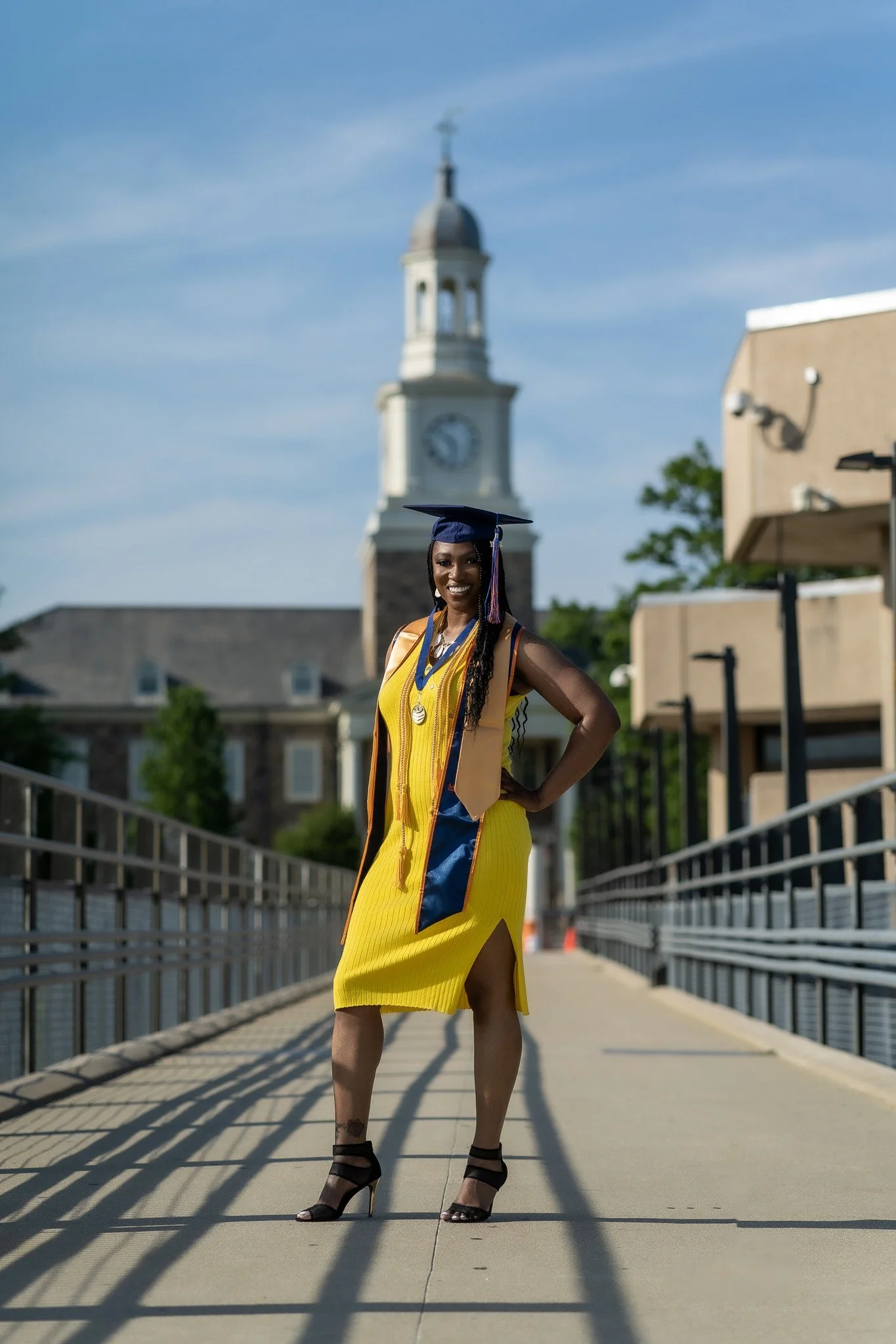 College graduate in a yellow dress, blue graduation cap and honor cords on a campus walkway