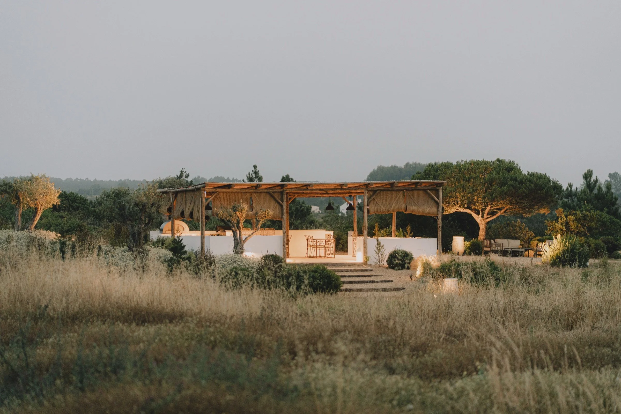 Open-air wooden pergola glowing at golden hour amid native gardens and olive trees at Amaria near Odeceixe