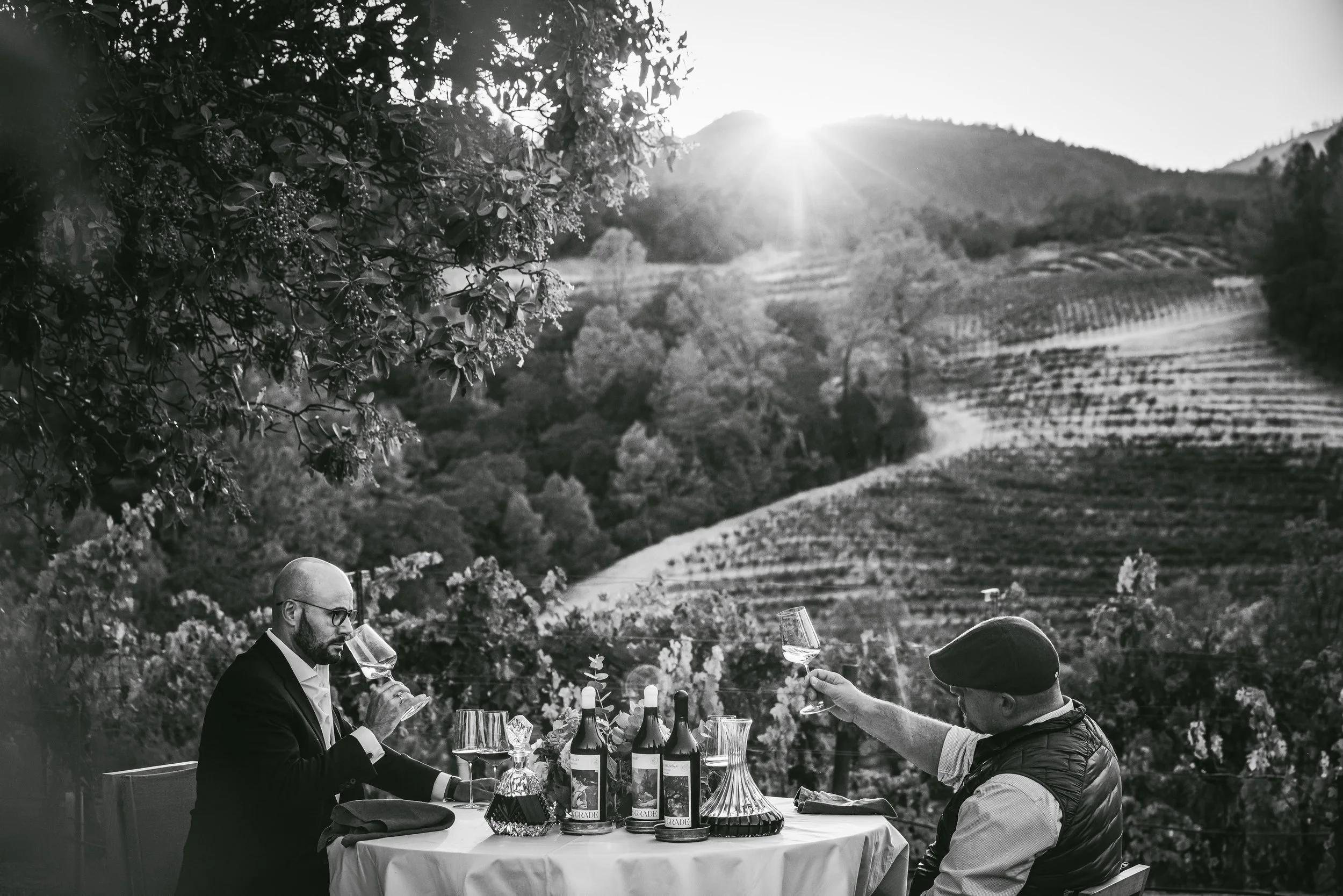 Two people sharing wine at a table beside a vineyard