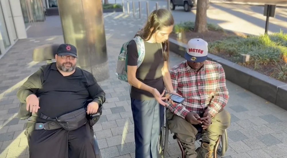 Three people outside a building, one using a wheelchair, look at an accessibility review on a phone