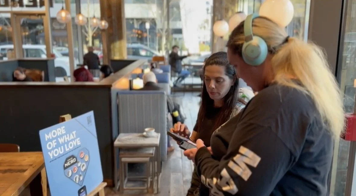 An intern wearing headphones reviews a café's accessibility on her phone while another person looks on