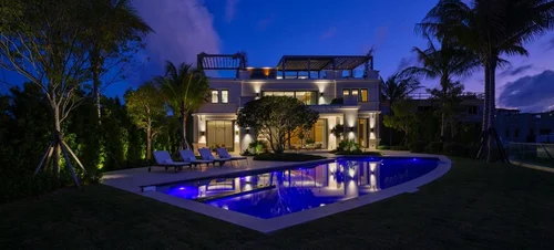 Vue nocturne de la villa avec piscine éclairée en bleu, transats, pelouse et palmiers