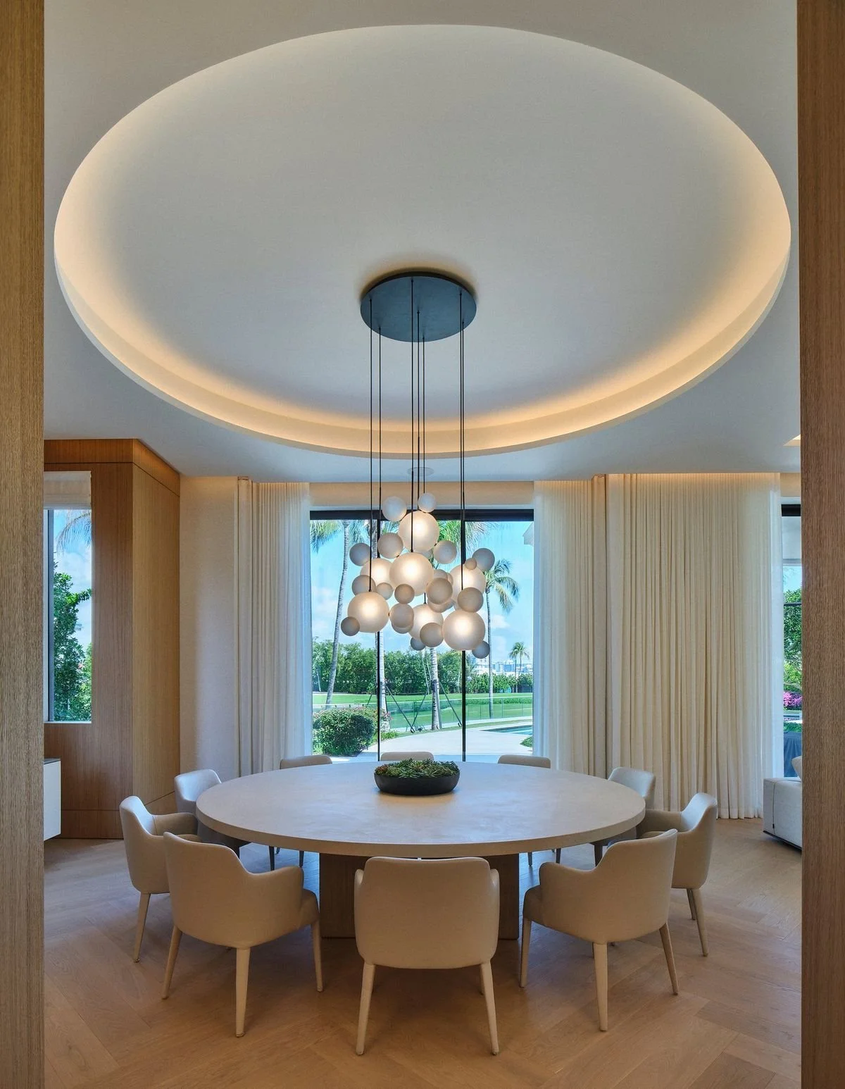 Salle à manger avec grande table ronde, chaises rembourrées, lustre à globes de verre et plafond circulaire éclairé