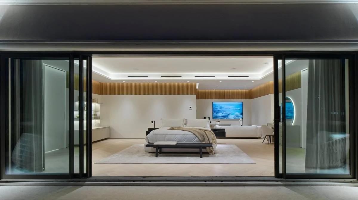 Chambre la nuit vue de la terrasse par les baies ouvertes, avec bandeau de tasseaux en bois et miroir rond éclairé