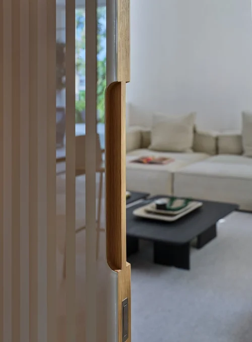 Porte au chant en bois avec poignée verticale encastrée, entrouverte sur un salon avec canapé clair et table basse sombre