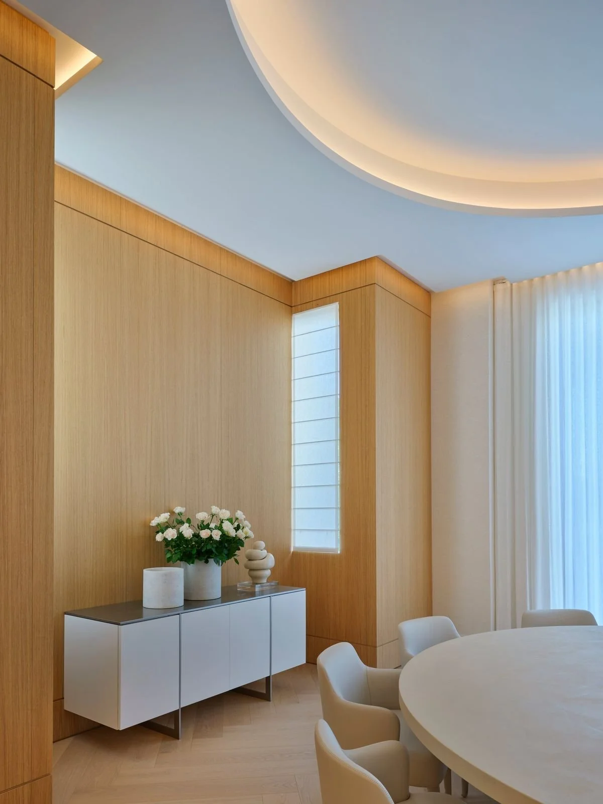 Coin repas avec boiserie, fenêtre verticale, enfilade blanche au plateau sombre, table ronde et plafond incurvé éclairé