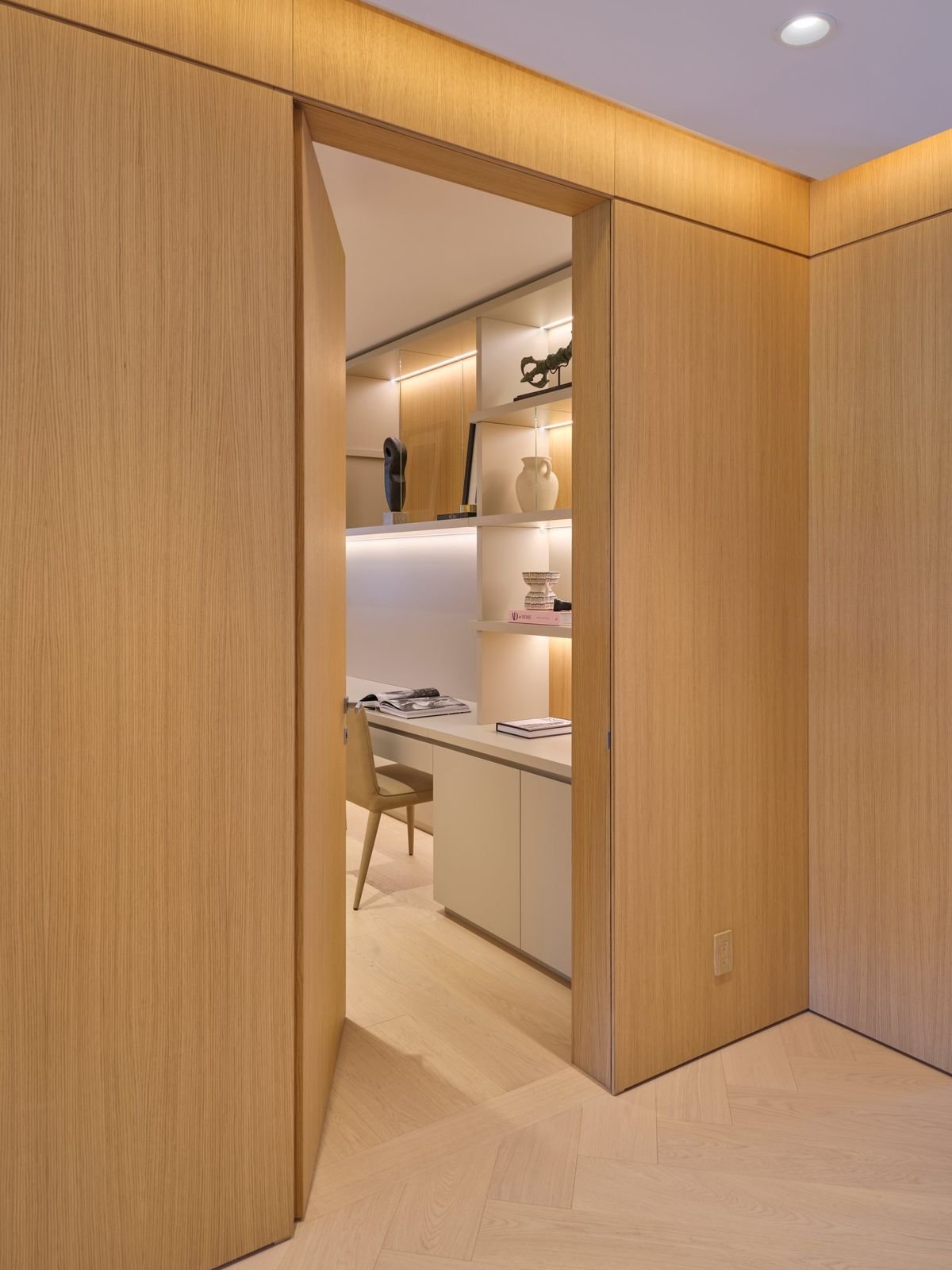 Haute porte habillée du même bois que la boiserie, ouverte sur un bureau avec étagères éclairées et plan de travail