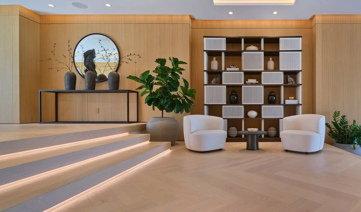 Salon avec boiserie, console et miroir rond, bibliothèque aux portes à lattes, deux fauteuils blancs et marches lumineuses