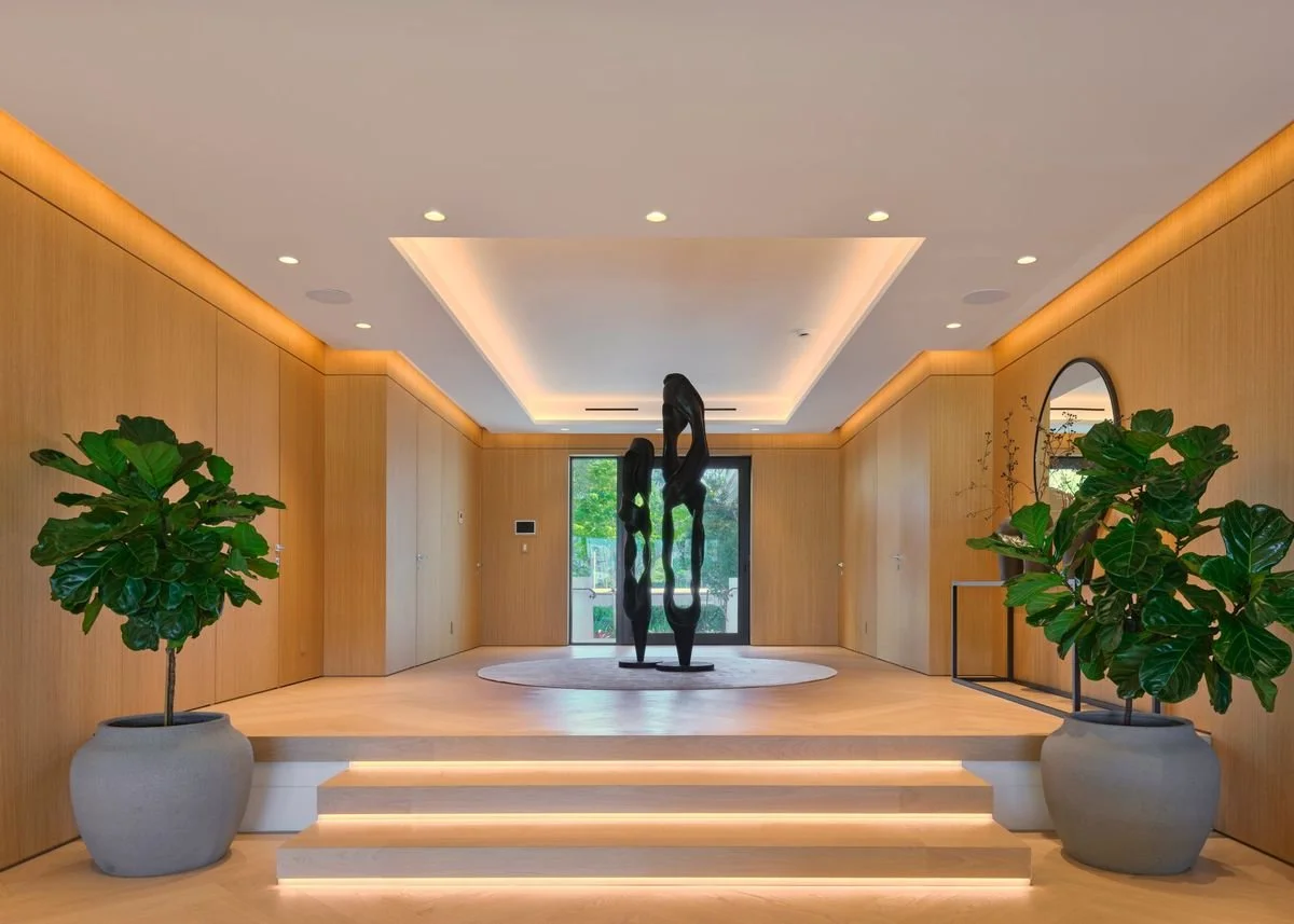 Hall d’entrée habillé de boiserie claire, marches lumineuses, plafond à éclairage indirect et porte vitrée sur le jardin