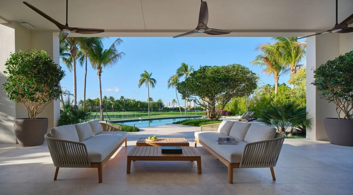 Loggia couverte avec canapés d’extérieur, tables basses et ventilateurs de plafond, ouverte sur piscine, palmiers et pelouse