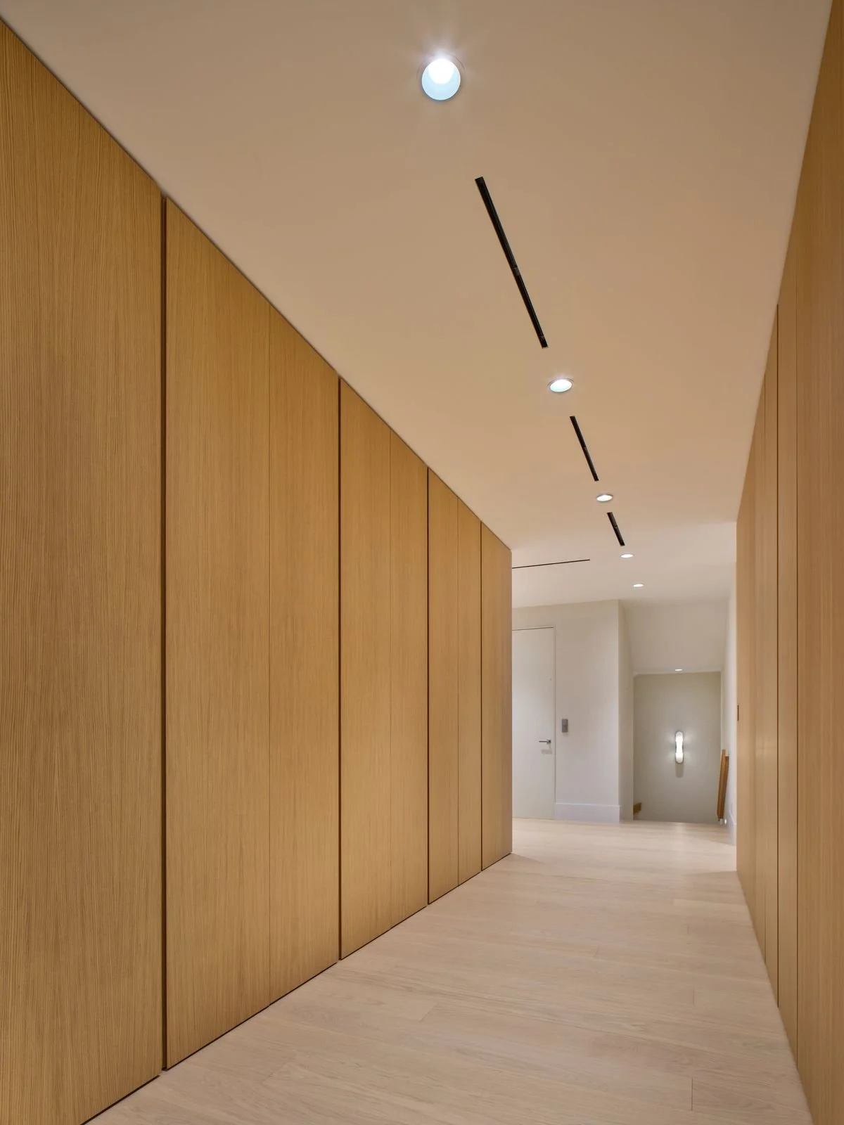 Couloir bordé d’armoires toute hauteur en bois clair aux portes sans poignées, avec luminaires linéaires au plafond