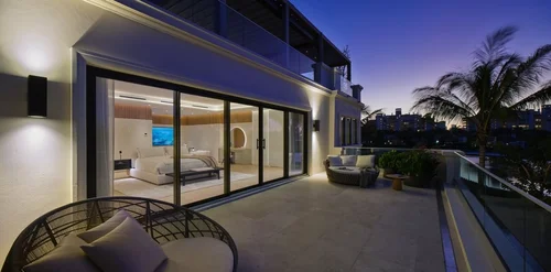 Terrasse au crépuscule avec assises d’extérieur, la chambre éclairée derrière les vitrages, palmiers en arrière-plan