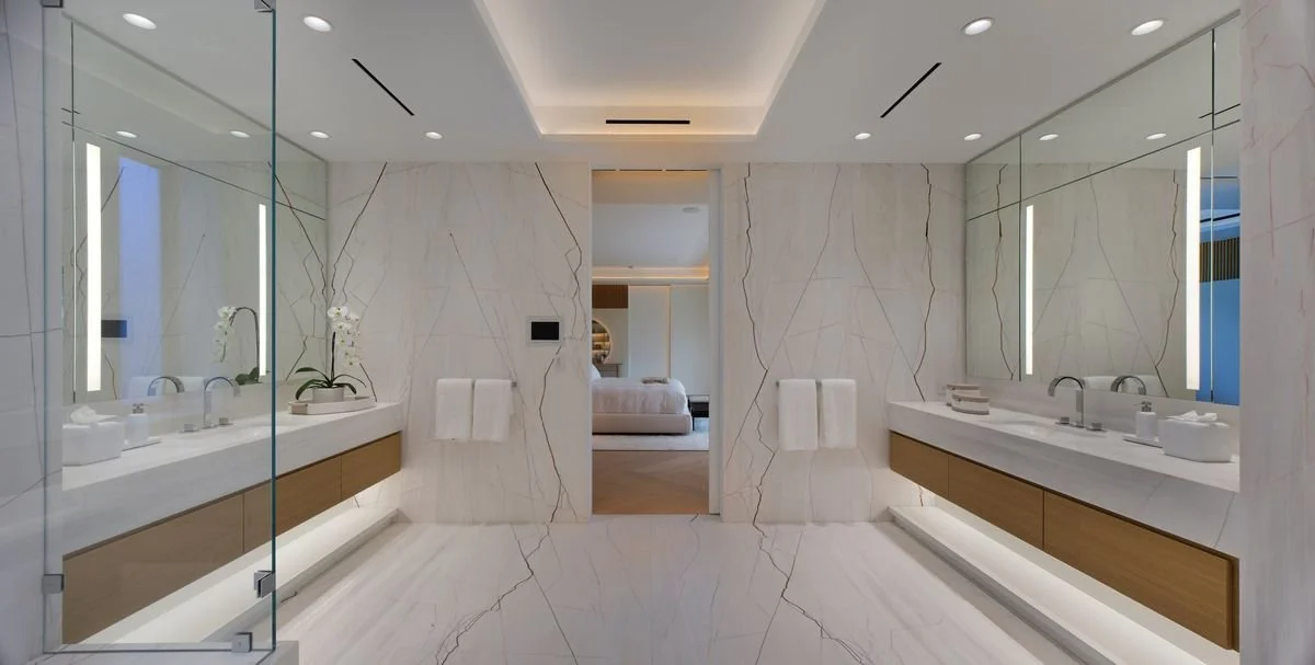 Salle de bains symétrique : deux meubles suspendus en bois et pierre, miroirs éclairés, murs en pierre veinée, douche vitrée