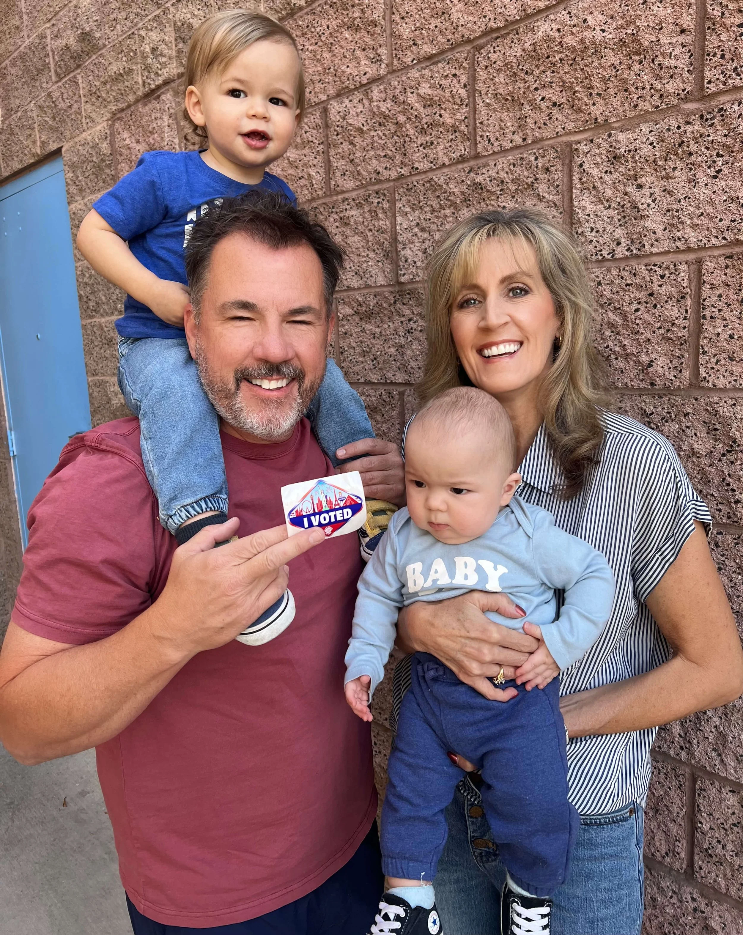 Melissa Hardy and her husband Damon holding their two young grandsons, one pointing to an I Voted sticker