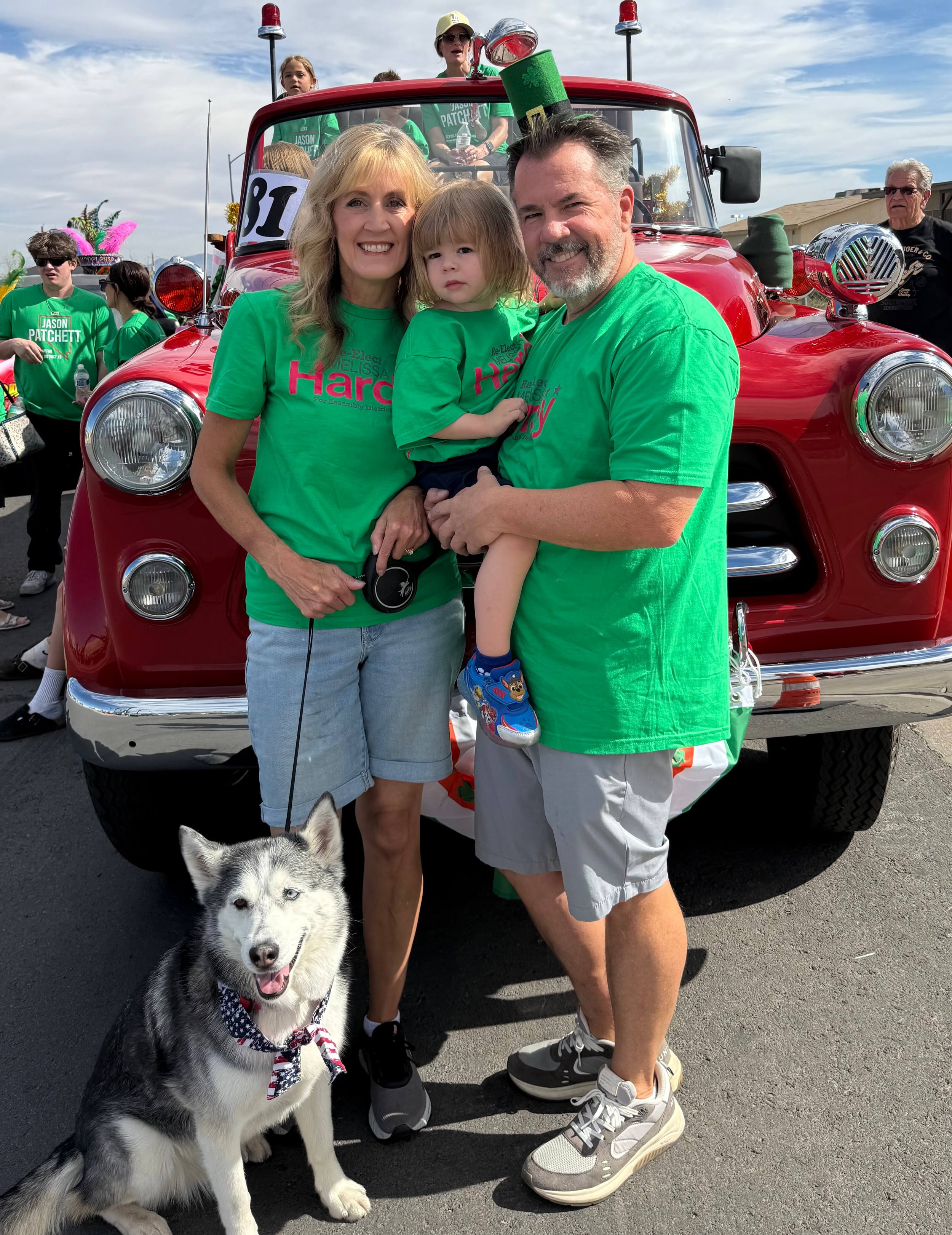 Melissa Hardy with her husband, a grandchild, and the family dog in front of a vintage red fire engine at a community parade