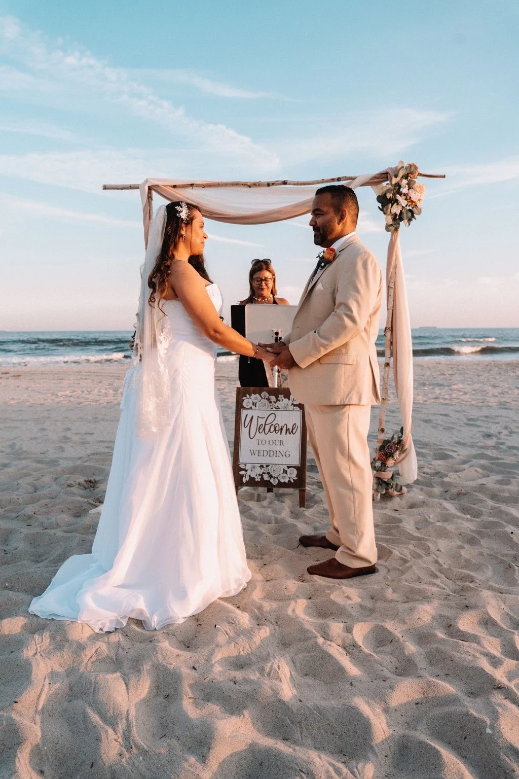 A couple holding hands under a beach wedding arch, with an officiant and welcome sign behind them.