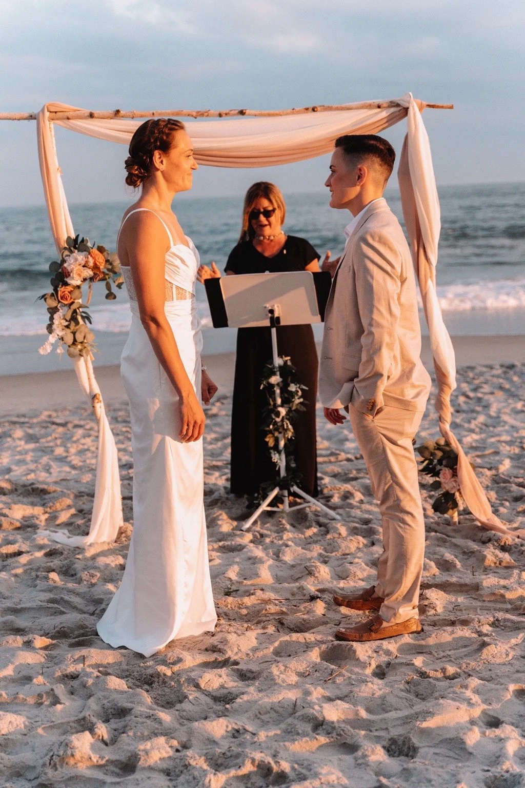 A couple facing one another beneath a draped beach wedding arch with their officiant behind them.