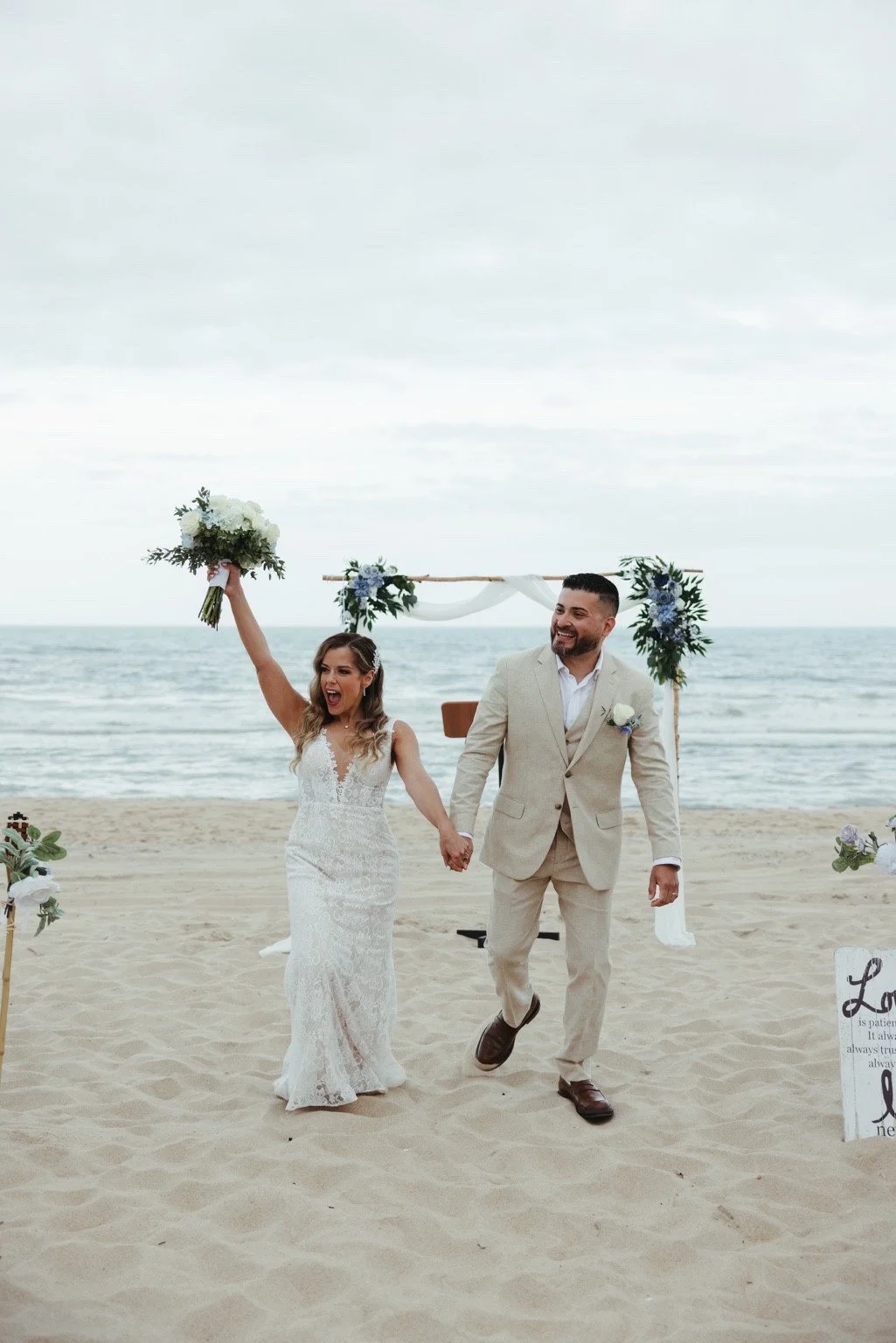 Newlyweds holding hands on a beach, with a bouquet raised and a floral arch behind them.