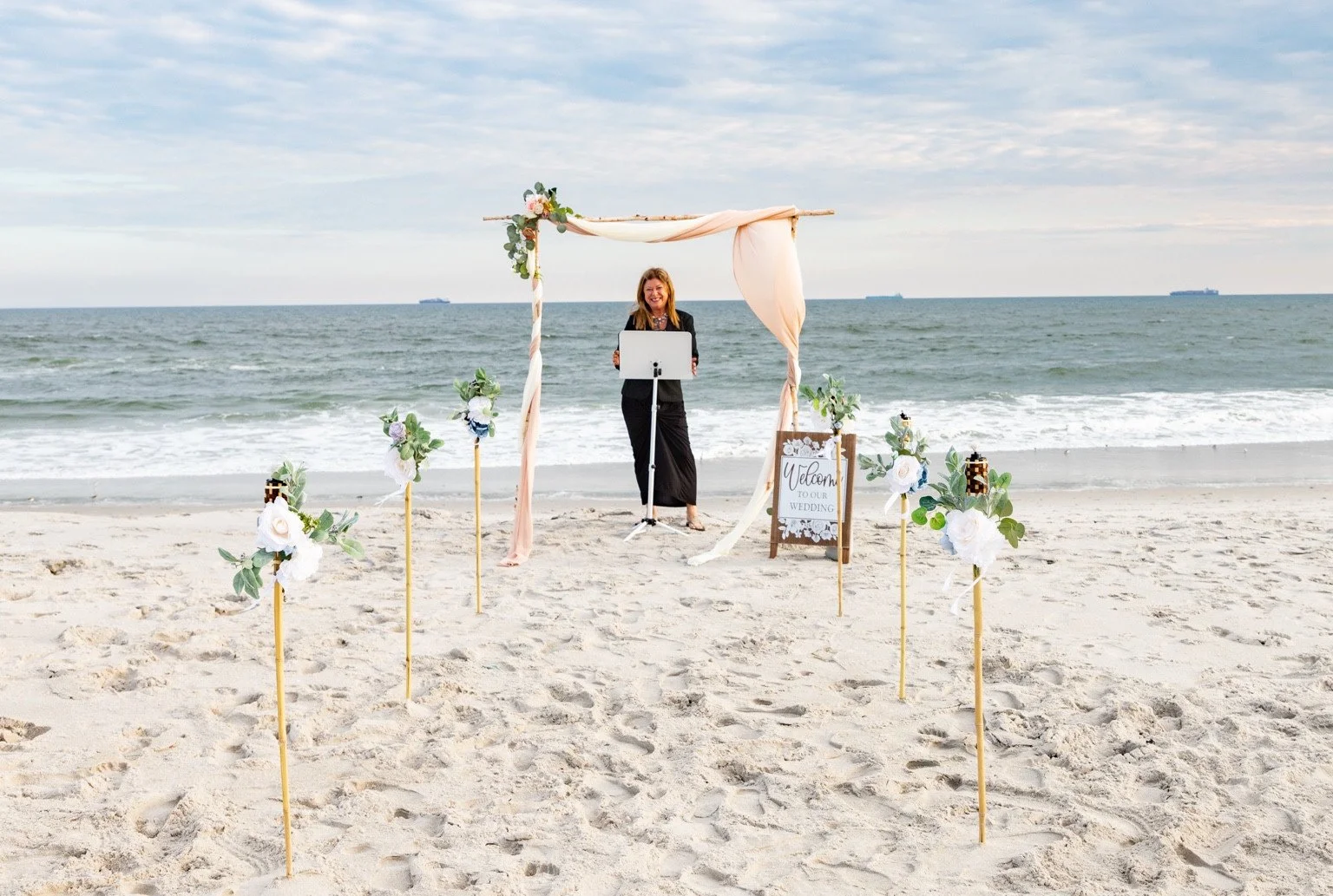 Wedding officiant standing beneath a draped beach ceremony arch, with floral aisle markers and the ocean behind.