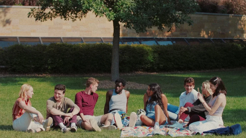 Eight friends sit together on a blanket on a sunlit campus lawn, talking beneath a tree