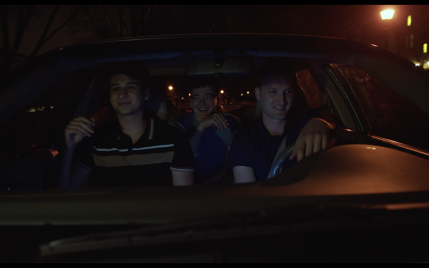 Three friends laugh together inside a car at night, street lights behind them