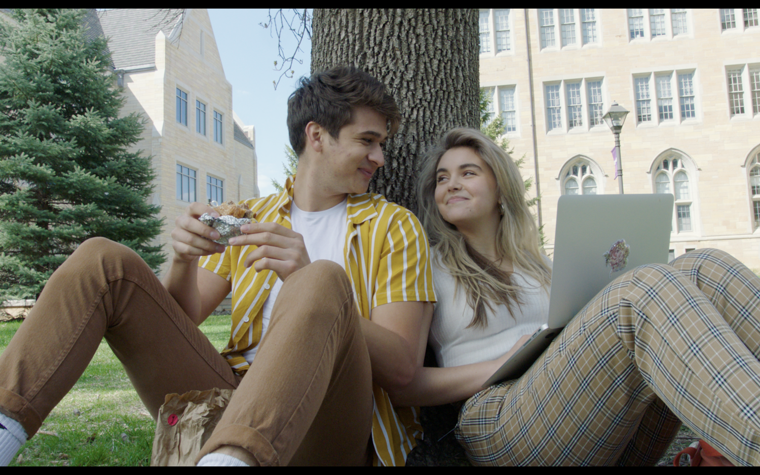 Two students sit against a tree on a campus lawn, one holding food, the other a laptop