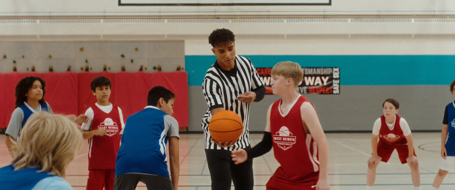 A referee holds the ball at centre court between two lines of youth players in red and blue jerseys