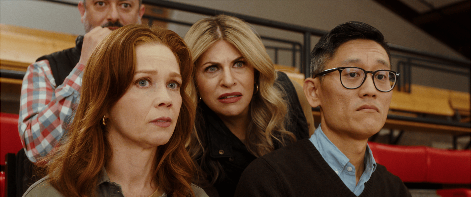 Three parents sit close together in the bleachers, watching the game with tense expressions