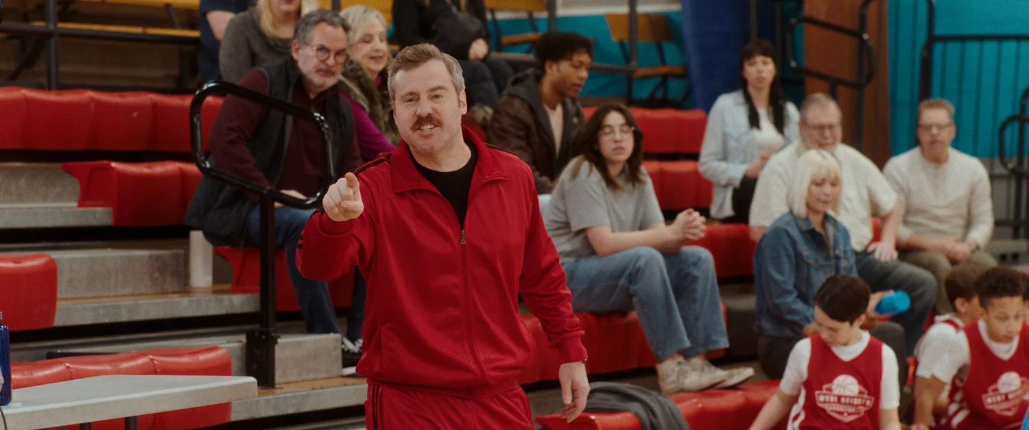 Tom stands at the sideline in a red tracksuit, pointing across the court, with parents watching from the bleachers behind him