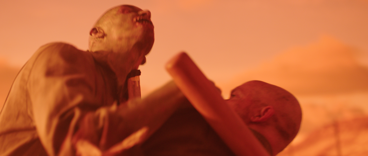 An actor jams a dull wooden board under the chin of an actor in zombie makeup