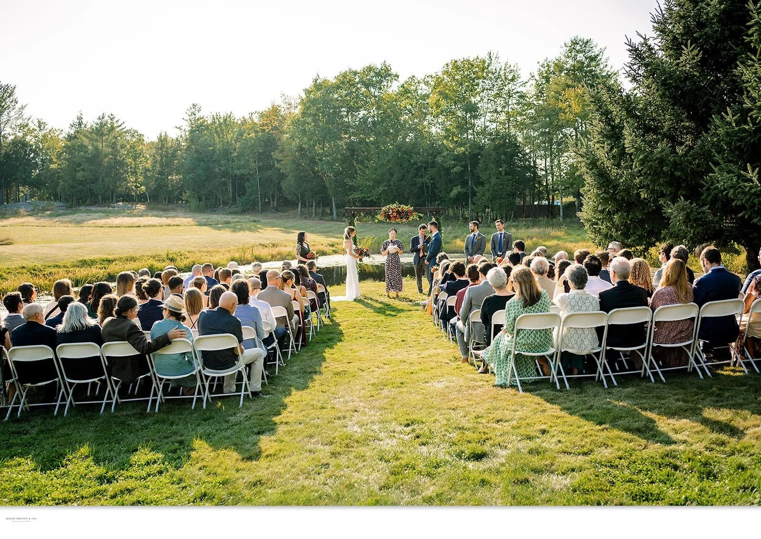 Guests seated for an outdoor ceremony on the lawn facing the pond