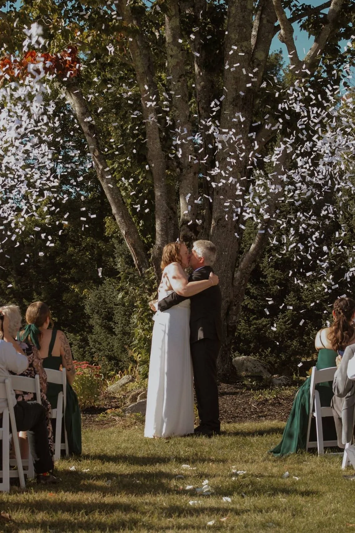 A couple sharing their first kiss under a big shade tree as confetti begins to fall above them at William Allen Farm