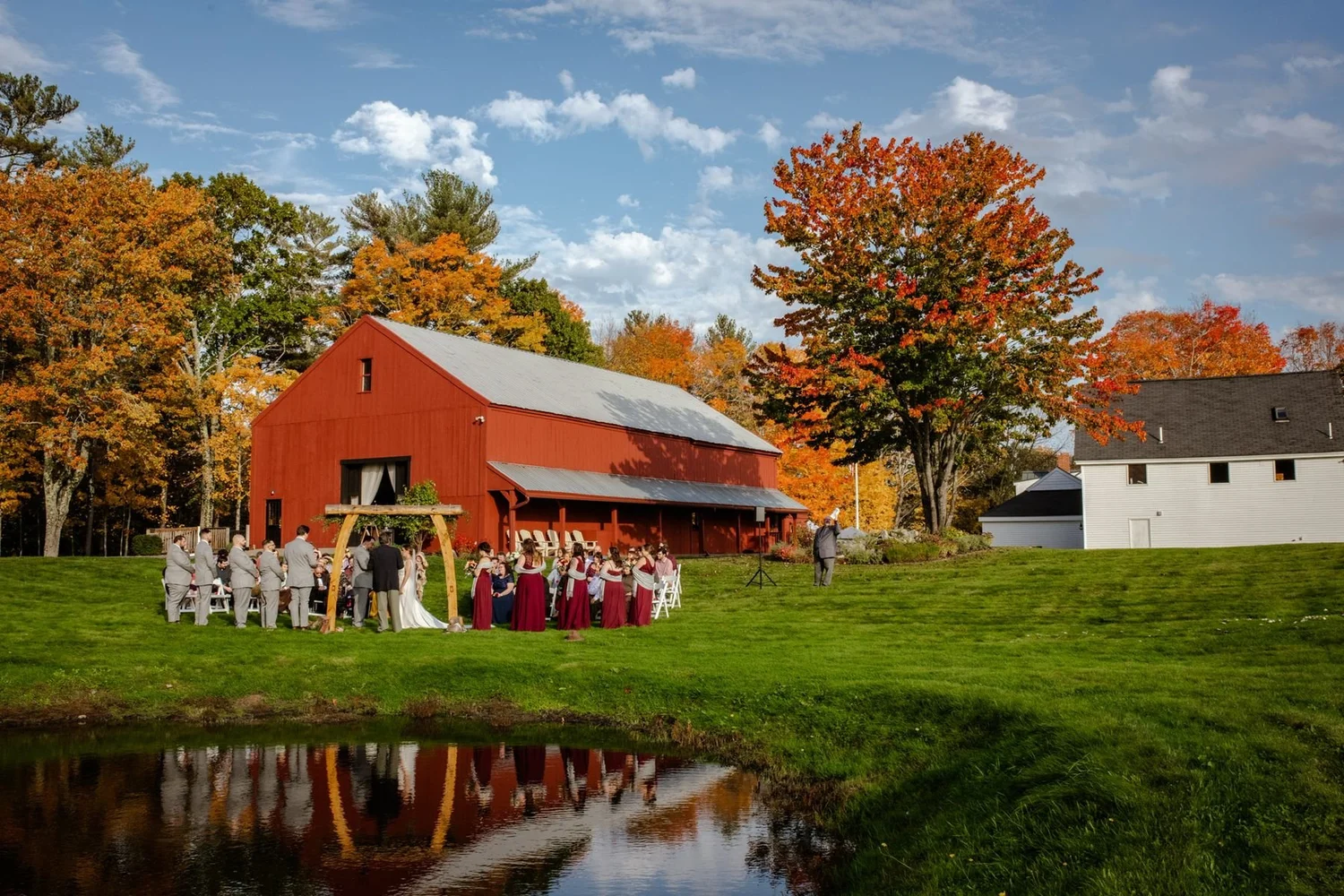 William Allen Farm's red timber-frame barn and pond in Pownal, Maine, with fall foliage and a ceremony set up by the water